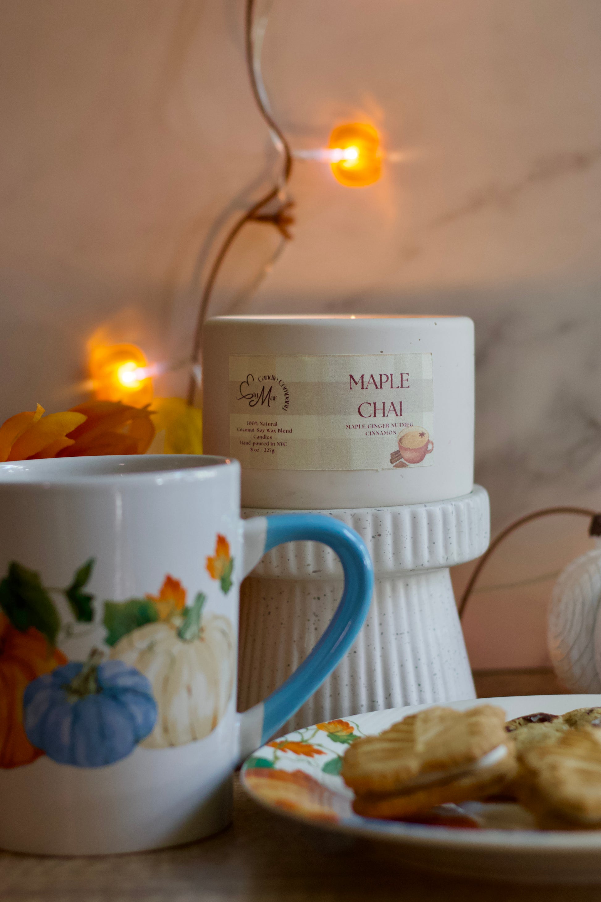 Cup with autumn-themed design, bowl of cookies, and candle on a decorative stand with warm lighting.